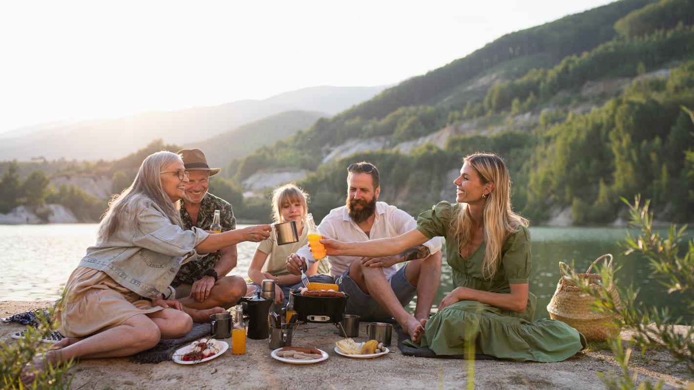 The image shows a diverse group of people sharing food by a serene lake, surrounded by nature. It conveys a harmonious and joyful atmosphere, highlighting health and wellbeing
