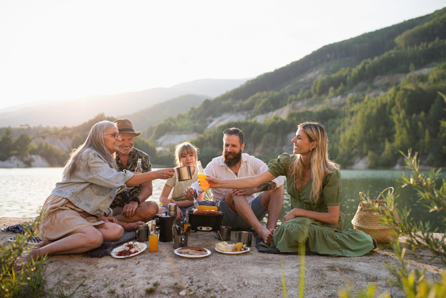 The image shows a diverse group of people sharing food by a serene lake, surrounded by nature. It conveys a harmonious and joyful atmosphere, highlighting health and wellbeing